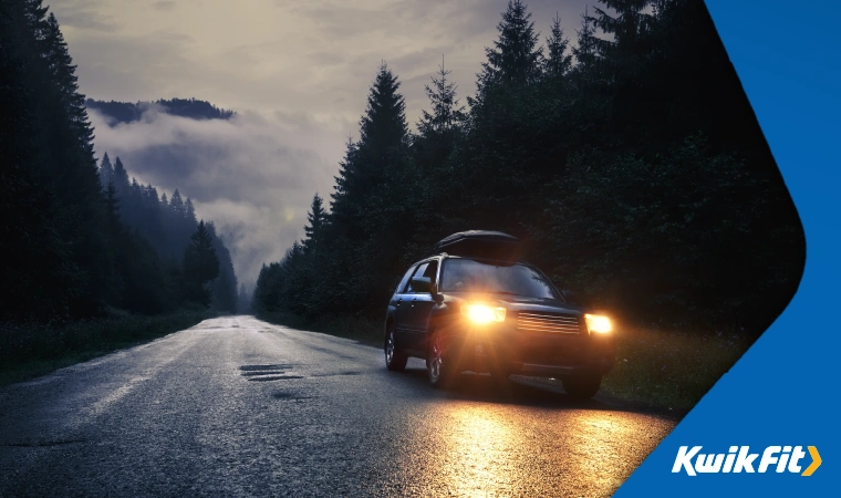 An SUV with its headlights on driving down a dark, tree-lined road.