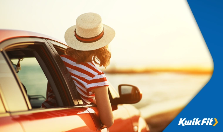 Woman with a red and white striped top and straw hat leans out of the front window of her red car that's parked near a beach.