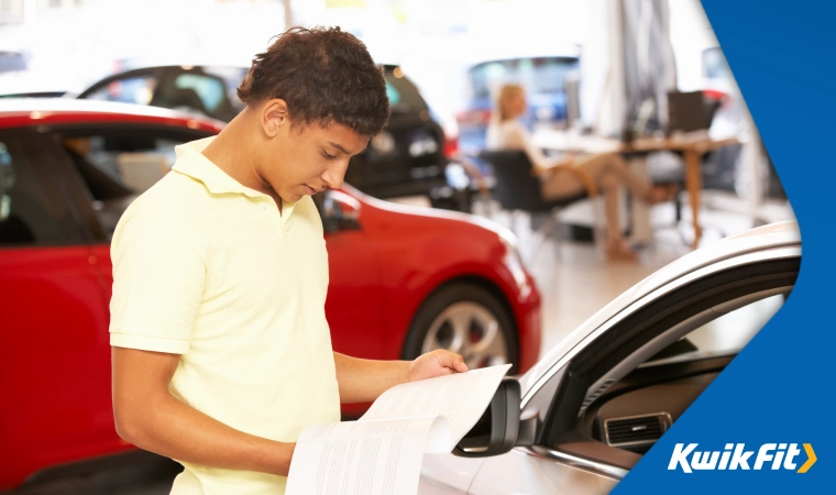 Person in a yellow polo shirt reading a brochure standing next to a car in a showroom.